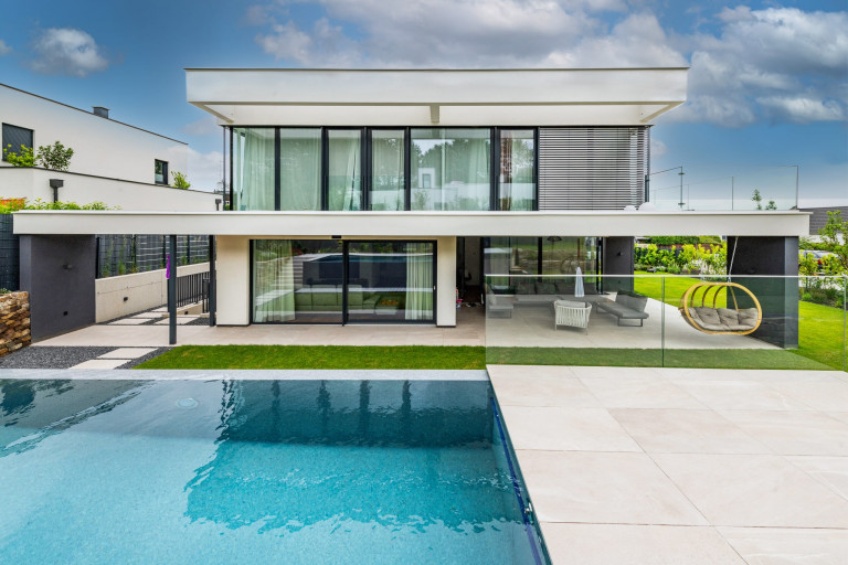 Modernes, helles Flachdach-Wohnhaus mit Glasgeländer-Balkon, großen Glasfronten, überdachter Terrasse und erhöhtem Pool von AL Architekt.