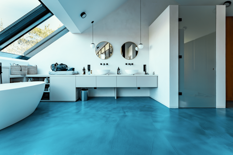 ARDEX präsentiert ein Badezimmer mit blauem Boden, einer weissen freistehenden Badewanne und einem Doppelwaschtisch mit runden Spiegeln von panDOMO®.