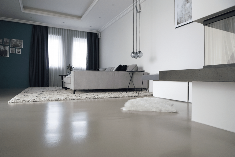 ARDEX zeigt ein Wohnzimmer mit einer großen, hellgrauen Couch auf einem hochflorigem Teppich, mit großen Fenstern, weissen Vorhängen und hellem Boden von panDOMO®.