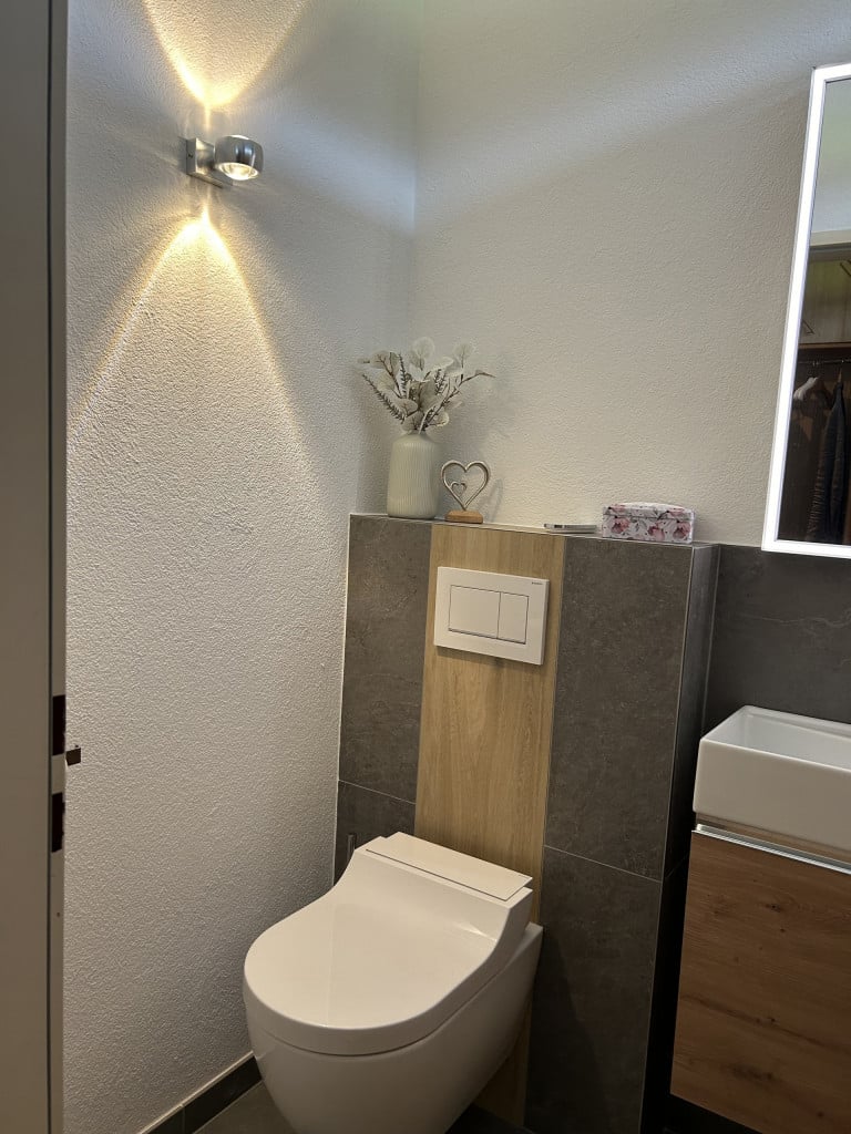 Badezimmer mit wandhängender Toilette, Wandleuchte, Spiegelschrank mit Rahmenbeleuchtung und schwebendem Doppelwaschtisch mit Stauraum in Holzoptik von der Bad Profi Salzburg Stadt.