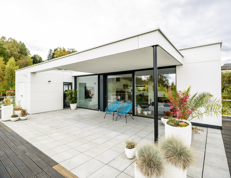 Bad Zeller zeigt die überdachte Terrasse eines weißen Hauses mit blauen Gartenstühlen, Pflanzen und hellgrauen Fliesen.