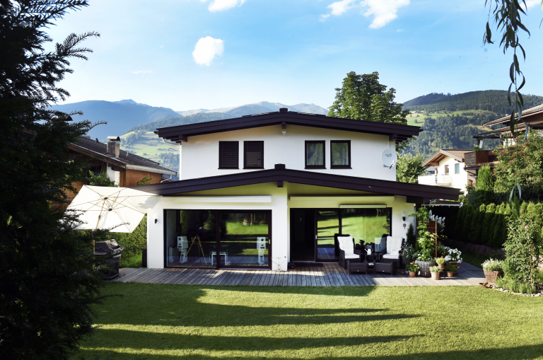 ERLER Haus zeigt ein weisses Einfamilienhaus mit zwei Stockwerken und einer überdachten Terrasse mit Holzboden und einer gemütlichen Sitzecke mit Rattanmöbeln und Glasschiebetüren mit Sicht auf den Wohnbereich.