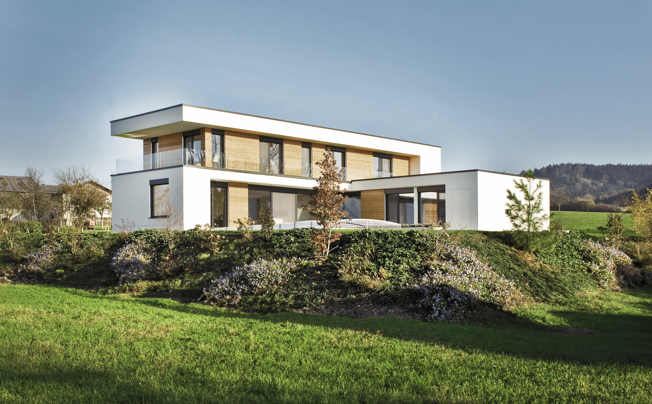 ERLER BAU GmbH zeigt ein modernes Haus mit Holzelementen und Flachdach, mit großen Fenstern und einem Balkon mit Glasgeländer, der sich um das ganze Gebäude erstreckt.