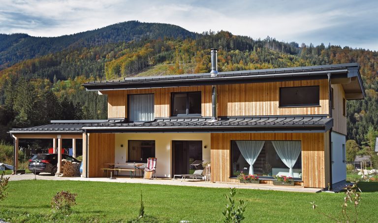 ERLER Haus zeigt ein großes Holzhaus im Grünen mit Carport und überdachter Terrasse mit einer Sitzecke.