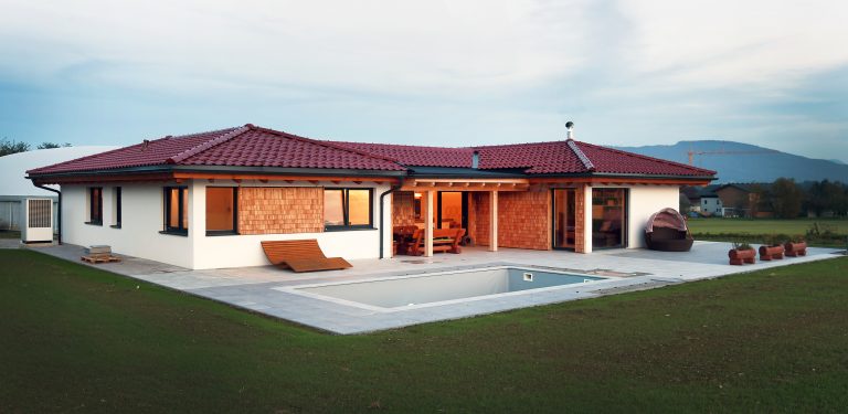 Ein schöner Bungalow mit rotem Dach, überdachter Terrasse mit einer Sitzgruppe aus Holz und großem Garten mit rechteckigem Pool von ERLER Haus.