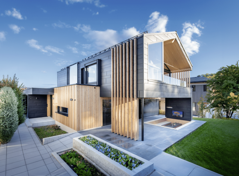 Erler Haus zeigt ein sehr modernes Holzriegelhaus mit gefliester Terrasse, dunkler Wandverkleidung, Balkon und eingebauter Sitzlounge inklusive Fernseher und Blumenbeeten.