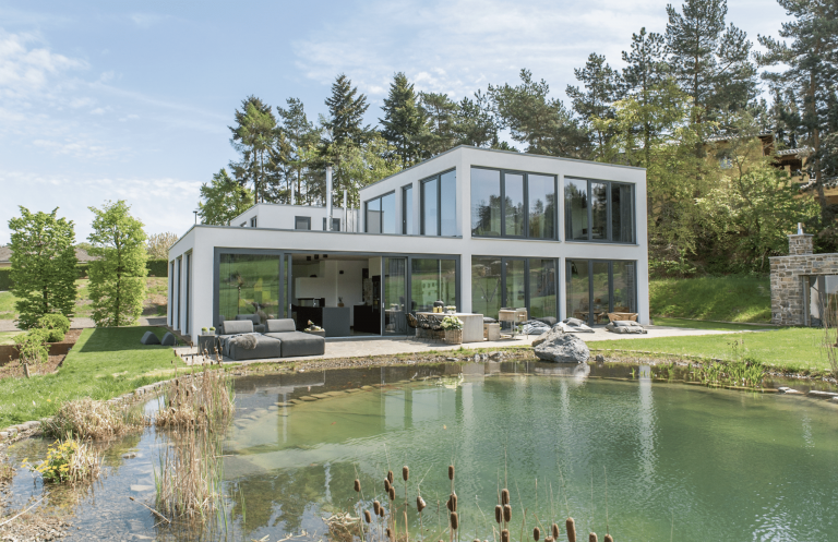 Modernes Flachdachhaus mit bodentiefen Holz-Alu-Fenstern von Gaulhofer mit großem Schwimmteich und gemütlichen Sitzmöbeln auf der Terrasse.