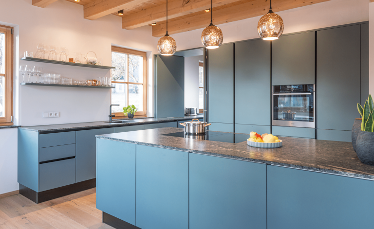 Moderne blaue Küche mit einer Arbeitsfläche aus dunklem Mamor, Holzdecke und hellen Wänden von WOHNHAUS Grill & Ronacher.