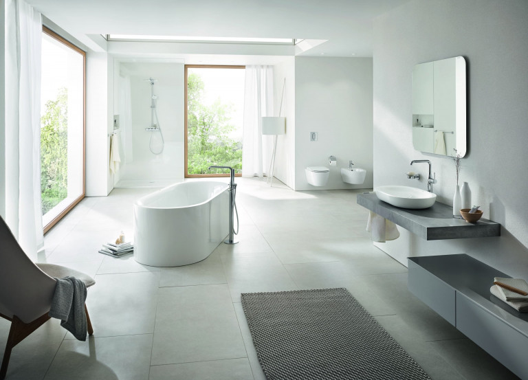 Badezimmer mit chrome Armaturen von Grohe mit weißer freistehender Badewanne, Regendusche und Glastrennwand, weißem Waschbecken mit breitem anthrazit Holzunterbau, weißem Wand-WC und Bidet und großflächigen beigen Bodenfliesen.