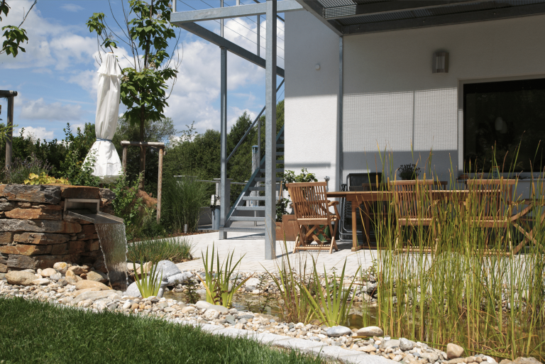 Hablesreiter zeigt eine überdachte Terrasse mit Gartenmöbel, einem kleinen Teich davor und ein integriertes Wasserspiel.