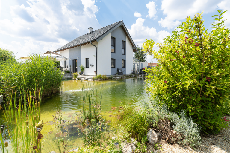 Weiß-graues eingeschößiges Haus von Hartlhaus mit großem Naturpool, wuchernden Pflanzen und einer einladenden Terrasse.