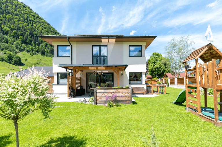 Hartl Haus zeigt ein helles Einfamilienhaus im Grünen, mit einem Spielplatz aus Holz und grüner Rutsche, einem Hochbeet und überdachter Terrasse.