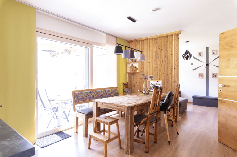 Hartl Haus zeigt ein Esszimmer mit gelben Wänden und Holztrennwand mit einem Holztisch und verschiedenen Stühlen vor einer großen Glastür.