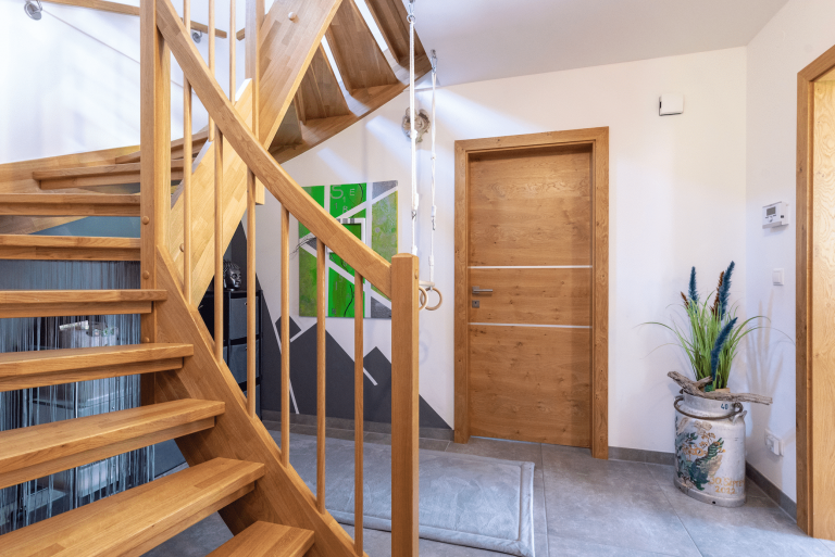 Hartl Haus zeigt einen Vorraum mit einem grauen Teppich, heller Wendeltreppe aus Holz, zwei Turnringen, einer Milchkanne mit Blumen und hellbraunen Holztüren.