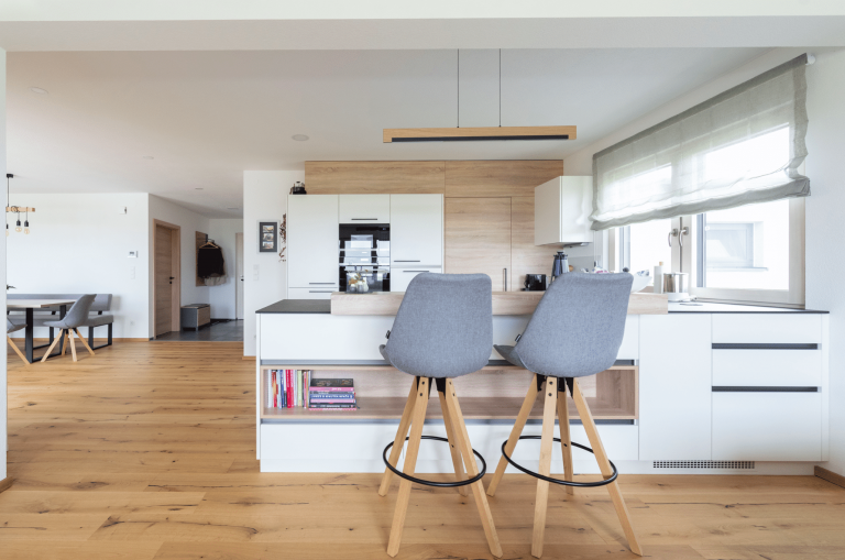 Hartl Haus zeigt eine große, helle Küche in L-Form mit weissen Oberflächen und Holzakzenten und grauen Hockern um an der Bar Platz zu nehmen.