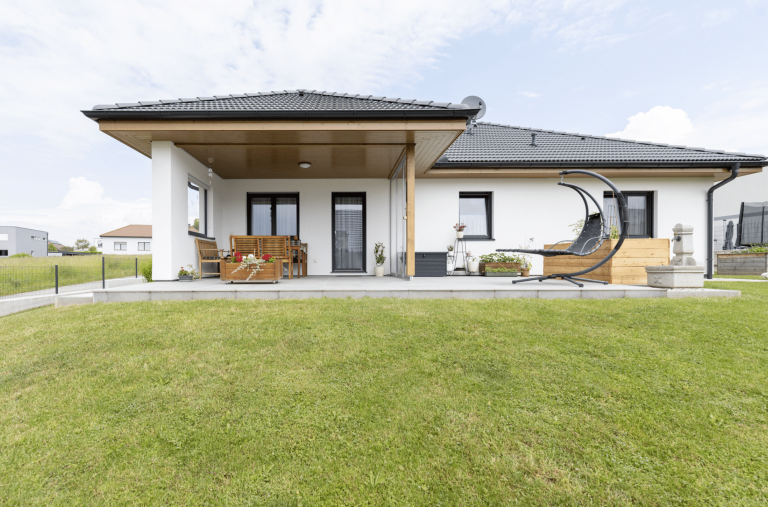 HARTL HAUS zeigt einen modernen, weißen Bungalow mit dunklem Dach, dunklen Türrahmen, großer Terrasse und verschiedene Outdoormöbel.