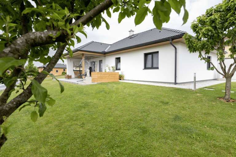 HARTL HAUS zeigt einen modernen, weißen Bungalow mit dunklem Dach, dunklen Türrahmen, großer Terrasse und verschiedene Outdoormöbel.