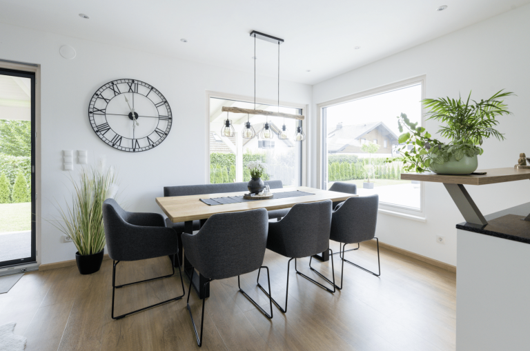 HARTL HAUS zeigt ein schön helles Esszimmer mit dunklen Stühlen, Holztisch, einer Uhr an der Wand und einer Hängelampe.