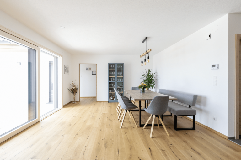 HARTL HAUS zeigt ein Esszimmer mit hellem Holzboden, grauen Polstermöbeln, einem Holztisch und blauer Glasvitrine.