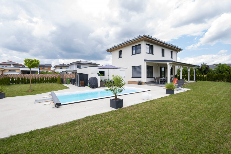 Einfamilienhaus mit flachem Satteldach, dunkelgraue Alufensterrahmen, überdachter Terrasse mit Stützsäulen und Pool von Hartl Haus.