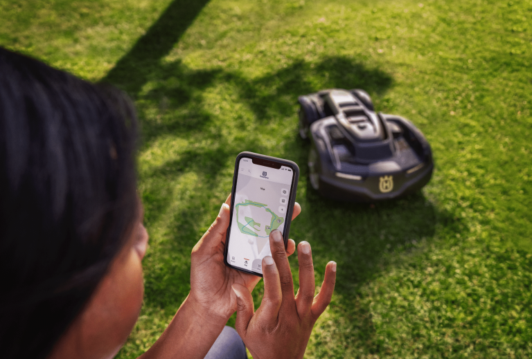 Husqvarna zeigt eine Frau, die den Mähroboter per App am Smartphone steuert.