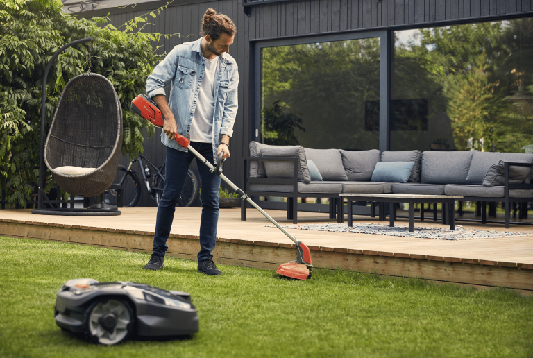 Husqvarna zeigt einen Mann, der mit Motorsense die Rasenkante mäht während der Mähroboter den Rasens pflegt.