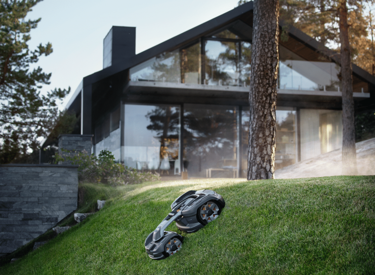 Husqvarna zeigt einen Mähroboter von Husqvarna, der einen steilen Hang im Garten eines Einfamilienhauses hinunter fährt.