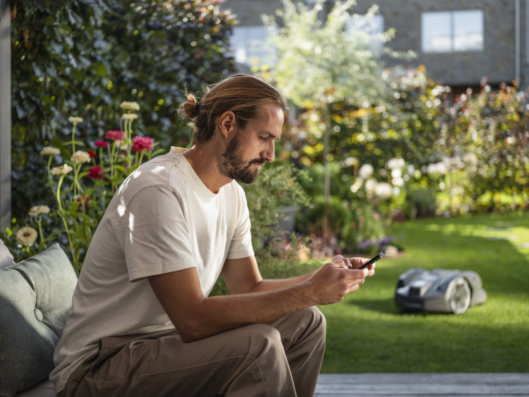 Ein Mann sitzt gemütlich auf seiner Terrasse und steuert über das Smartphone den Automower 305E NERA von Husqvarna.