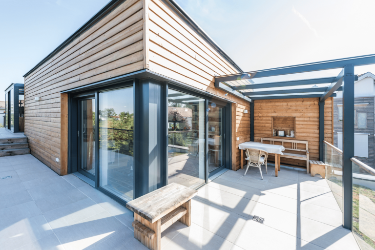 Terrasse mit Natursteinplatten, Holz-Gartenmöbeln, teilweise Überdachung aus Glas sowie deckenhohen Fenstern und Schiebe-Terrassentüren von Internorm.