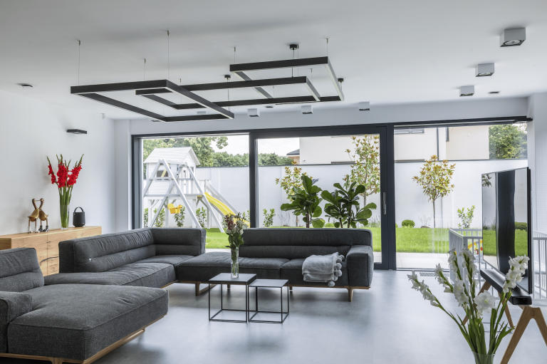 Internorm zeigt ein Wohnzimmer mit dunkelgrauer Couchlandschaft und moderner Lampe in schwarz mit Schiebetür zum Garten und dem Spielplatz mit Rutsche.