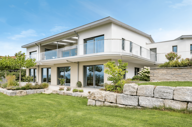 Internorm zeigt ein modernes Haus mit Flachdach und großem Balkon mit Glasgeländer und Glastüren im Erdgeschoß mit Zugang zur Terrasse und dem gepflegten Garten.