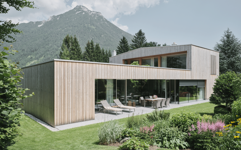 Josko zeigt ein modernes Einfamilienhaus mit Flachdach, einer hellen Holzverkleidung und sehr großen Glasfronten und einer Schiebetür aus Glas zur überdachten Terrasse.