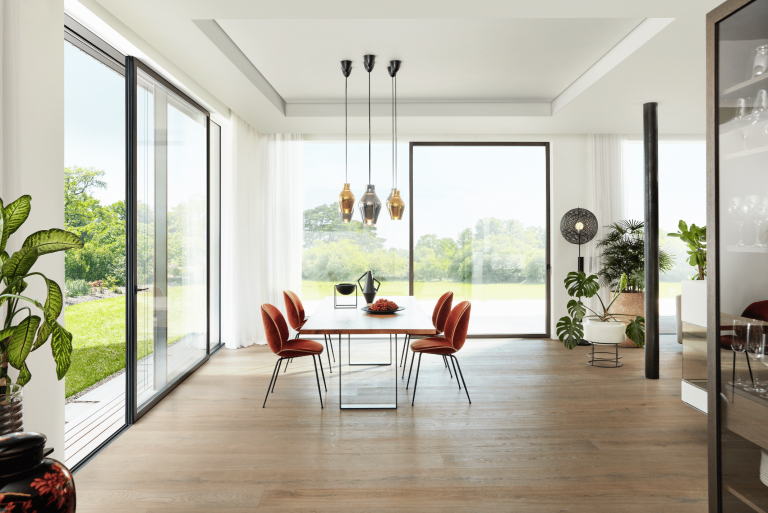 Josko zeigt ein helles Esszimmer mit Holzboden und einem eckigen Tisch mit orangenen Stühlen und großen Glasfronten und einer Glastür mit Zugang in den Garten.