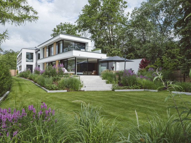 Moderne weiße Flachdachvilla mit Ganzglassystem und Glasschiebetür mit schwarzem Rahmen von Josko vor dem Stiegenabgang in den Garten und Terrasse mit Glasgeländer.