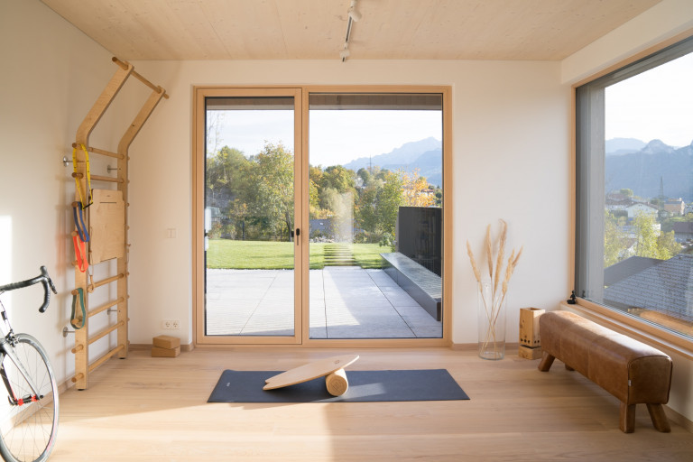 Heller Fitnessraum mit großem fix verglasten Fenster, Glasfront und Glastüre mit hellbraunem Rahmen von Josko mit Blick auf Terrasse und Garten.