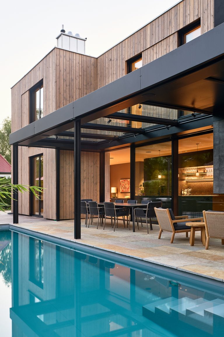 Modernes Flachdach-Holzhaus mit Glas überdachter Terrasse, Ganzglassystem und Glasschiebetüre mit schwarzem Rahmen von Josko mit vorgelagerter Terrasse mit Pool.