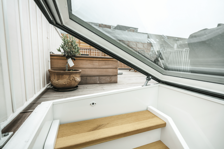 Lamilux zeigt den Ausstieg zu einer Dachterrasse durch ein großes Fenster mit schwarzem Rahmen und einer Holztreppe.