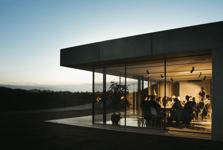 Moderner Flachdach-Bungalow bei Abendstimmung mit Holzterrasse, bodentiefer Fixverglasung und Hebeschiebetür von M SORA mit Bilck in das Esszimmer.