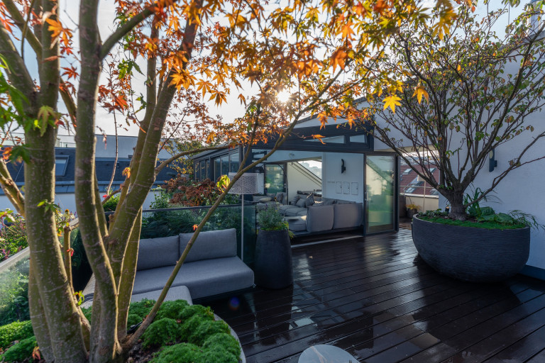 Praskac Pflanzenland Prachtgarten zeigt Dachterrasse mit Wintergarten, Outdoor-sitzbank und Pflanzentrögen mit Laubäumen.