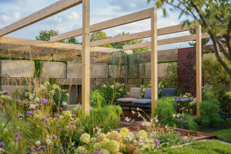 Praskac Pflanzenland Prachtgarten zeigt Blumenbeete und Sträucher vor einer Pergola mit Terrassenboden, Sitzbank und Steinmauer.