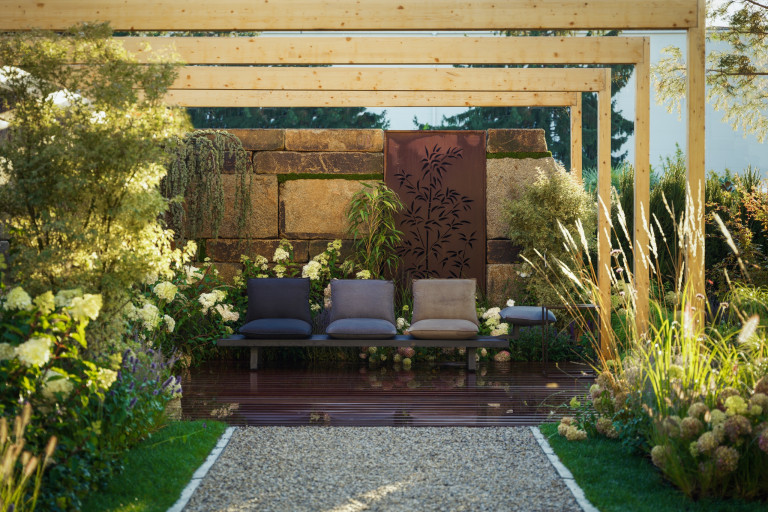 Praskac Pflanzenland Prachtgarten zeigt Schotterweg der zu einer Pergola mit Terrassenboden, Sitzbank und Steinmauer führt gesäumt mit Blumenbeeten und Sträuchern.