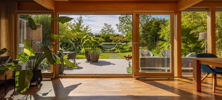 Praskac Pflanzenland Prachtgarten zeigt Wohnraum mit Blick auf Terrasse und Garten mit Pflanzentrögen, Bäumen und Sträuchern und Steinplatten im Rasen.