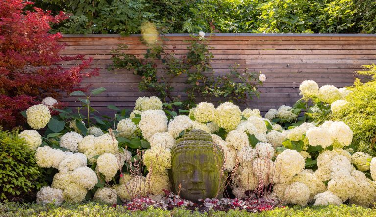 Praskac Pflanzenland Prachtgarten zeigt eine Beet mit Bodendeckern, Sträuchern und einer Skulptur vor einem Holzzaun.