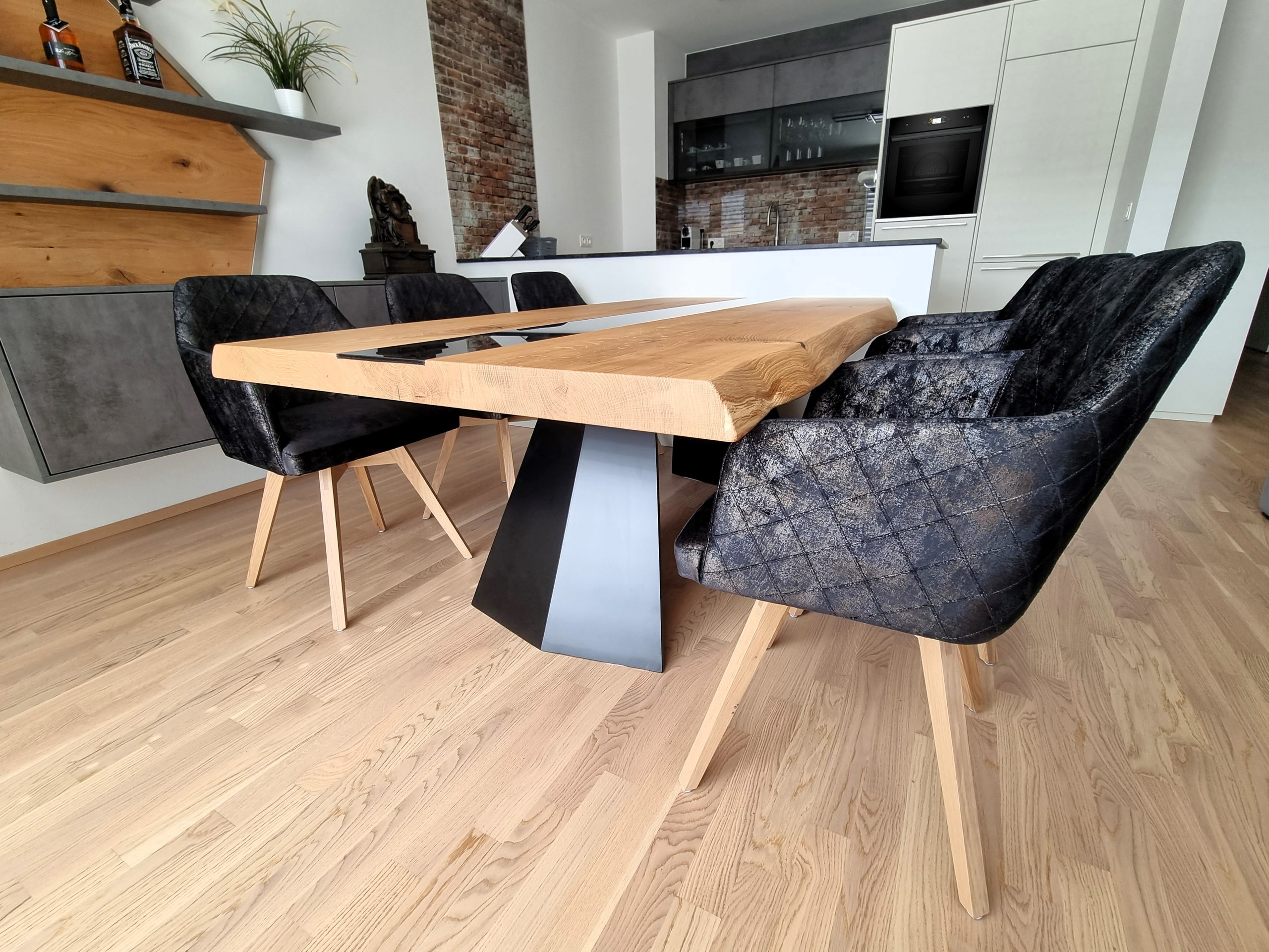 Küche mit Holzesstisch mit schwarzem Glaselement und schwarzen Tischfüßen, Polsterstühlen in schwarzem Used-Look mit Metallic-Elementen. einem schwebenden Sideboard mit Wandpaneel in Betonoptik und Holz und einer Inselküche in einem Materialmix aus Weiß, Betonoptik und Ziegelwandoptik von Pröll Wohnideen.