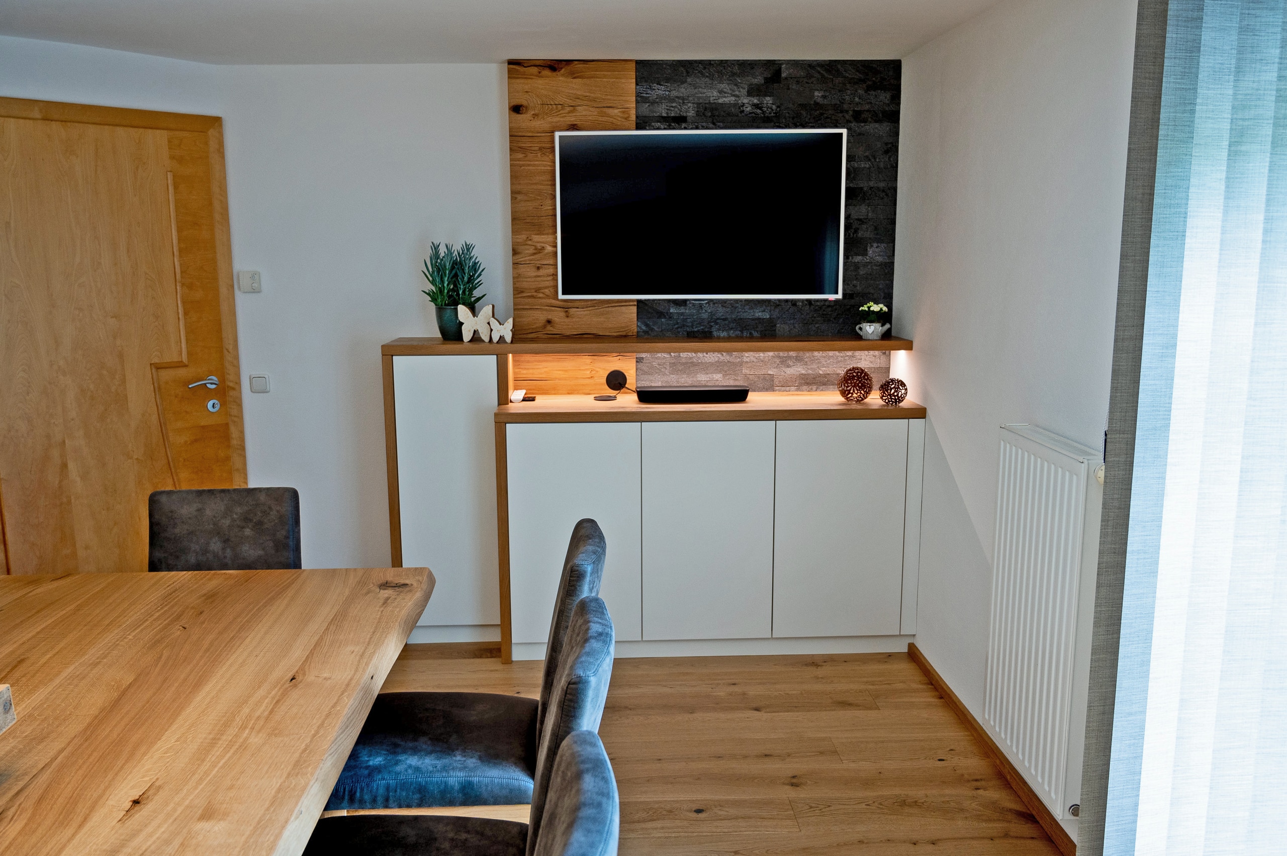 Wohnraum mit Esstisch in Holz, Polsterstühlen und einem indirekt beleuchteten Medienregal mit Wandpaneel und Flatscreen von Pröll Wohnideen.