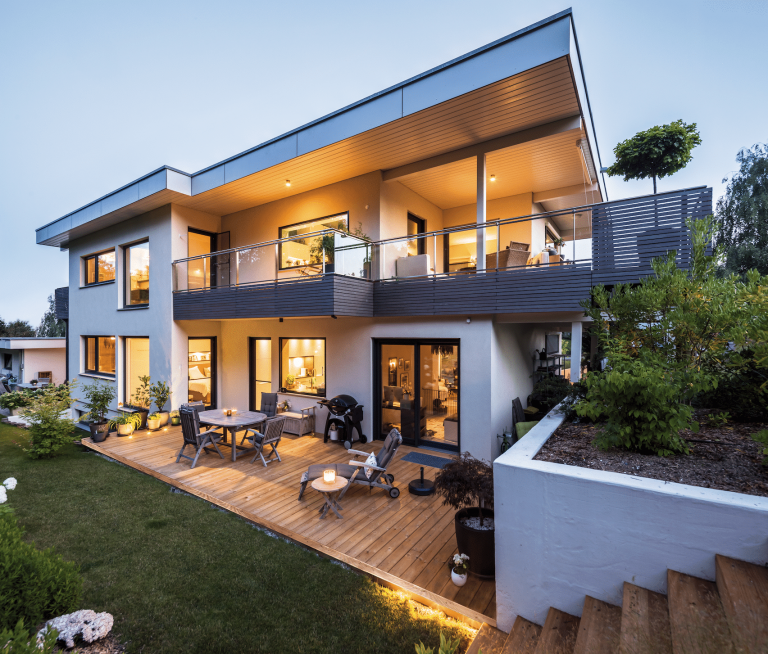 Regnauer Fertighaus zeigt ein modernes Einfamilienhaus mit Flachdach und Balkon mit Glasgeländer und Holzverkleidung, großer Terrasse bei Abenddämmerung.