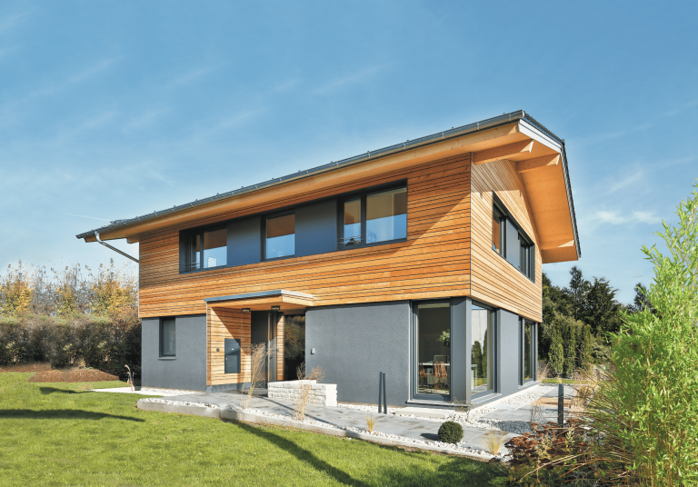 Regnauer zeigt ein graues Einfamilienhaus mit Holzverbau im oberen Bereich, großen Fenstern, einem dunklen Dach und gepflegtem Garten.