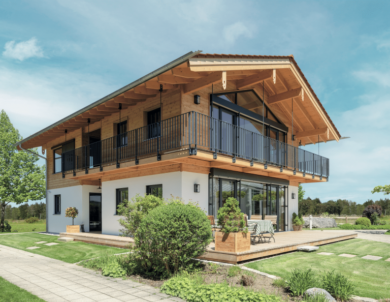 Regnauer zeigt ein Einfamilienhaus mit Holzverkleidung, durchgehendem Balkon mit Glasfront und schwarzem Geländer und eine Terrasse mit Essgruppe.