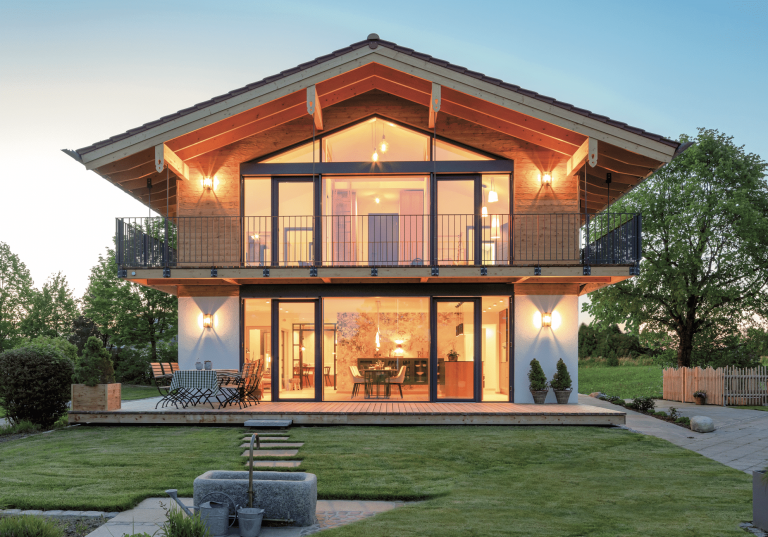 Regnauer zeigt ein Einfamilienhaus aus Holz mit großen Glasfronten und einem durchgehendem Balkon, einer Terrasse und viel Beleuchtung in der Abendstimmung.