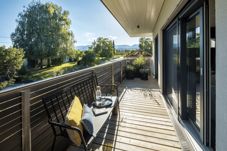 Regnauer zeigt einen langen Balkon mit Holzboden und einer gemütlichen Sitzbank mit dekorativen Kissen und großen Pflanzen in schwarzen Töpfen.